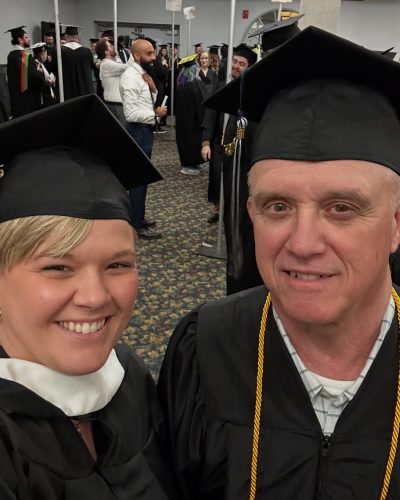 Christine Knight and father Sean McCormick, both dressed in caps and gowns smile for a the picture on UMA graduation day 2025. Fellow graduates and helpers in the background.