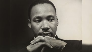Black-and-white portrait of a young MLK Jr in a suit and tie, hands clasped beneath his chin, looking downward and to the side with a thoughtful, reflective expression.