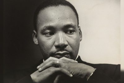 Black-and-white portrait of a young MLK Jr in a suit and tie, hands clasped beneath his chin, looking downward and to the side with a thoughtful, reflective expression.