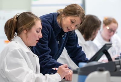 A UMA instructor leans over a lab bench to guide a student in a white lab coat as they work with laboratory equipment during a laboratory sciences class.