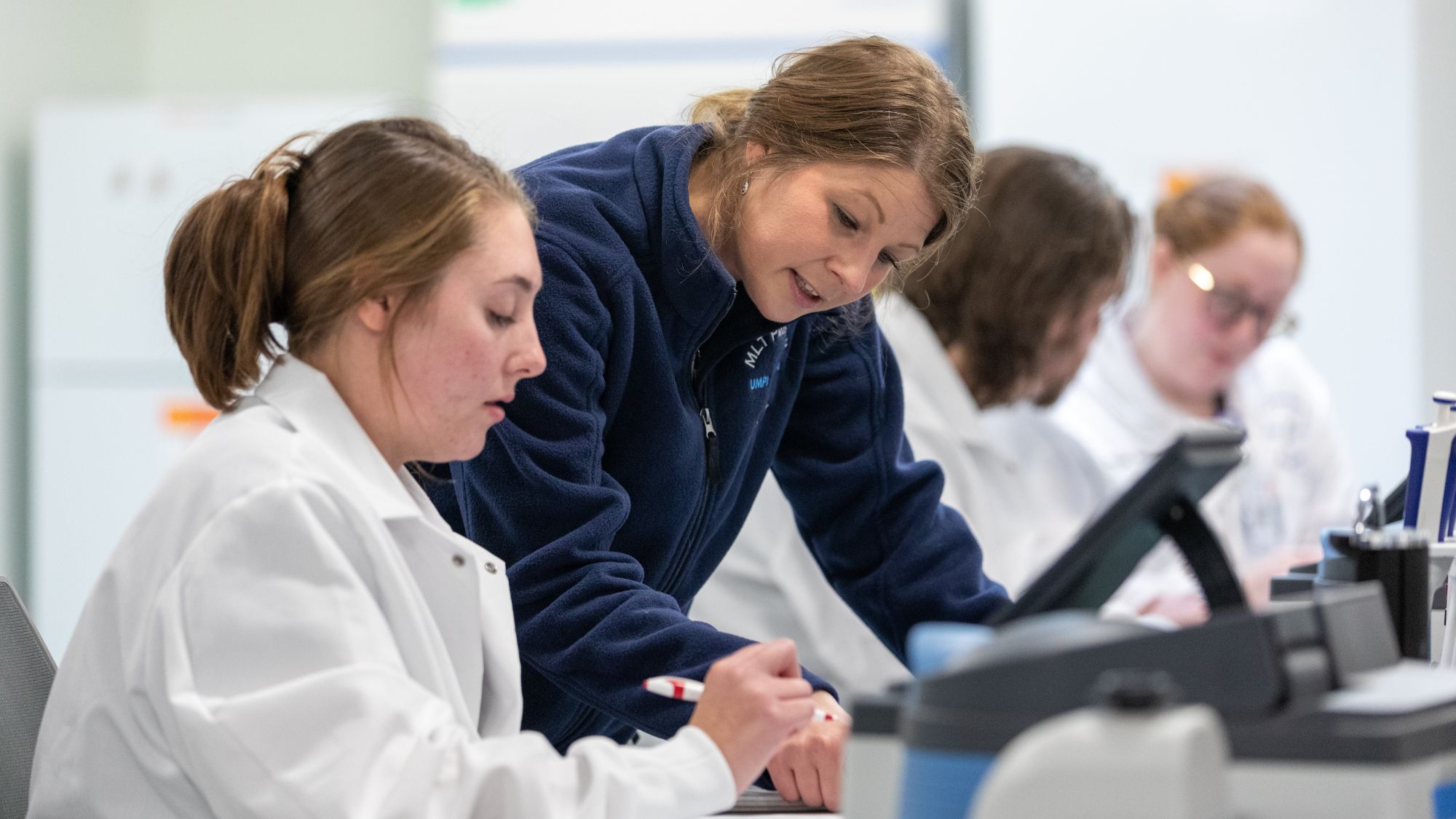 A UMA instructor leans over a lab bench to guide a student in a white lab coat as they work with laboratory equipment during a laboratory sciences class.