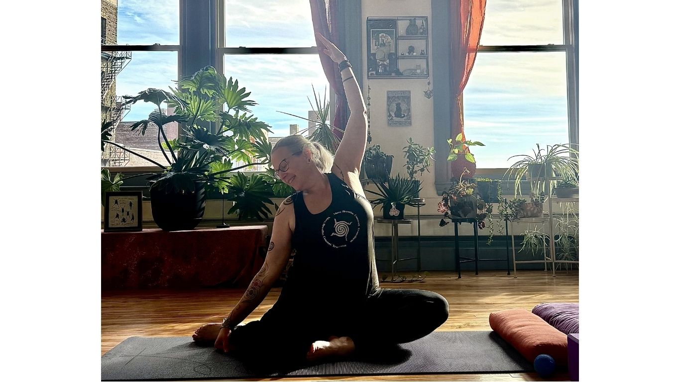A person sits on a yoga mat in a sunlit room filled with plants, gently stretching one arm overhead in a seated pose. The space feels calm and welcoming, reflecting a relaxed, trauma-informed yoga practice.