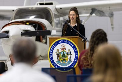 Isabella Puleo speaks to an audience in an aircraft hangar, with a plane visible behind her, during the Aviation Maintenance School Open House event.