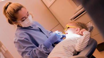 A dental hygienist wearing a mask, gloves, and safety glasses examines a young child’s teeth. The child reclines in a dental chair wearing protective glasses while the hygienist uses a dental tool under an overhead light.