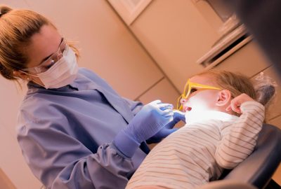 A dental hygienist wearing a mask, gloves, and safety glasses examines a young child’s teeth. The child reclines in a dental chair wearing protective glasses while the hygienist uses a dental tool under an overhead light.