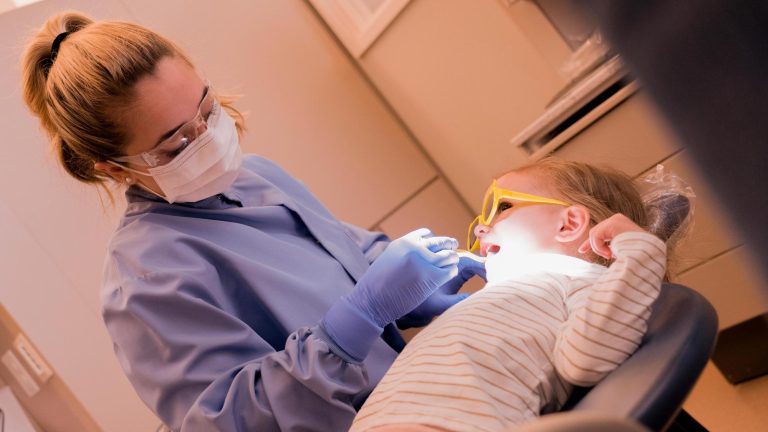 A dental hygienist wearing a mask, gloves, and safety glasses examines a young child’s teeth. The child reclines in a dental chair wearing protective glasses while the hygienist uses a dental tool under an overhead light.