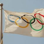 white flag that shows the five colroed olympic rings, flying in the breeze in front of a blue sky.