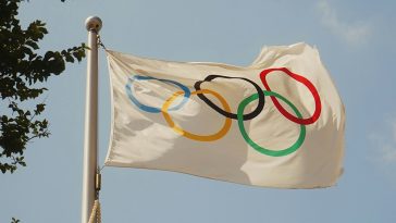 white flag that shows the five colroed olympic rings, flying in the breeze in front of a blue sky.