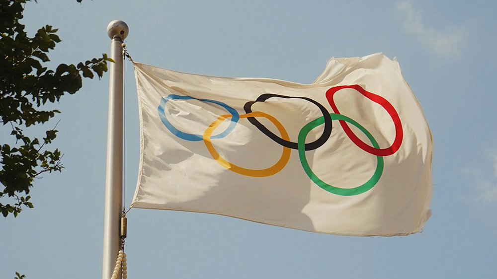 white flag that shows the five colroed olympic rings, flying in the breeze in front of a blue sky.