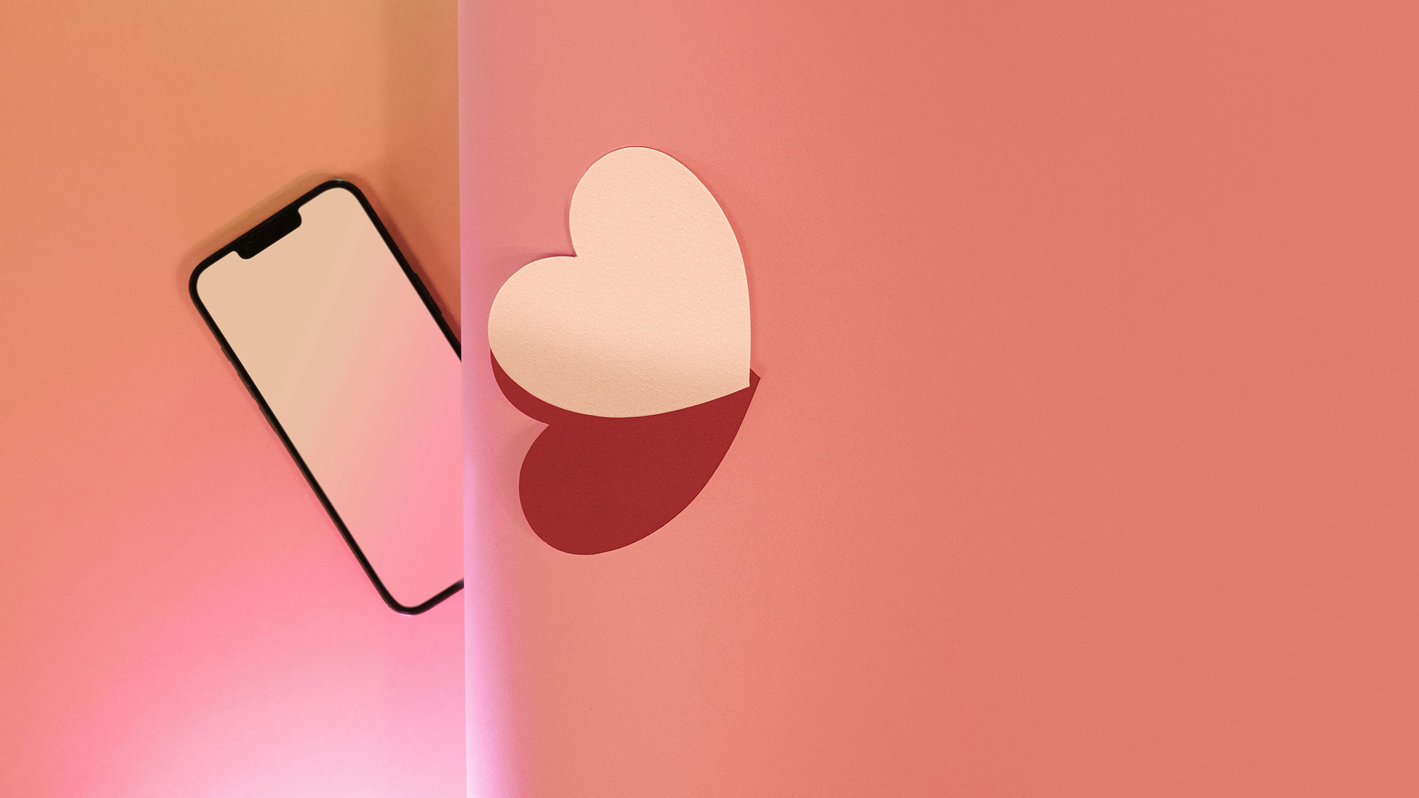 A smartphone lies on a pink gradient background, partially separated by a vertical shadow from a white heart shape on the right,