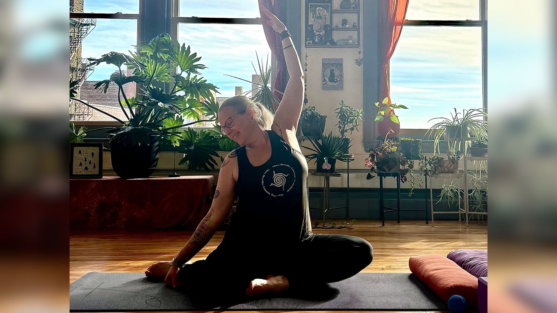 A person sits on a yoga mat in a sunlit room filled with plants, gently stretching one arm overhead in a seated pose. The space feels calm and welcoming, reflecting a relaxed, trauma-informed yoga practice.
