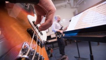 Close-up of a person’s hand plucking the strings of a bass guitar in the foreground, with another musician and a music stand holding sheet music slightly out of focus in the background during a rehearsal.