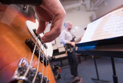 Close-up of a person’s hand plucking the strings of a bass guitar in the foreground, with another musician and a music stand holding sheet music slightly out of focus in the background during a rehearsal.