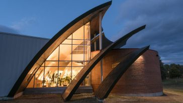 Exterior of the Michael Klahr Center at the University of Maine at Augusta at dusk, showing a brick cylindrical wing and several large, curved wooden and metal “petal” structures framing a tall glass façade that glows with warm interior light, symbolizing renewal and reflection.