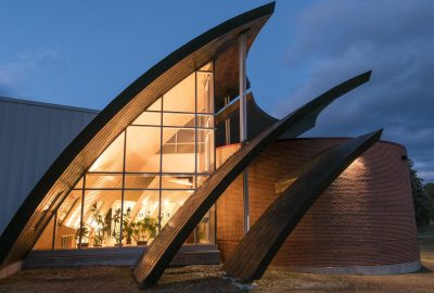Exterior of the Michael Klahr Center at the University of Maine at Augusta at dusk, showing a brick cylindrical wing and several large, curved wooden and metal “petal” structures framing a tall glass façade that glows with warm interior light, symbolizing renewal and reflection.