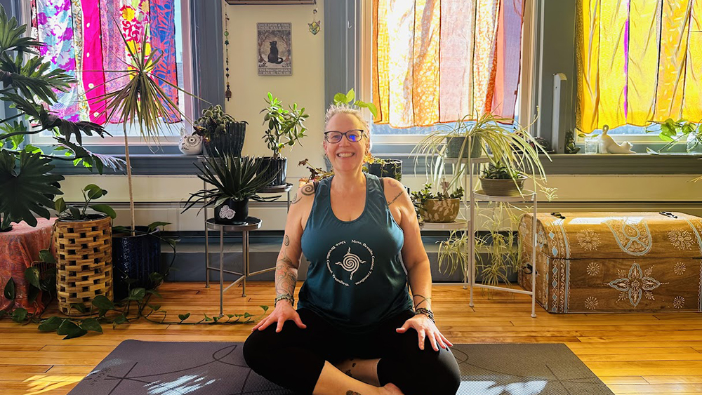 Sarah Hentges sits cross-legged on a yoga mat in a bright, plant-filled room, smiling at the camera. She wears glasses, a green tank top, and black leggings. Sunlight filters through colorful patterned curtains behind her, and numerous potted plants surround the space, creating a warm, inviting atmosphere.