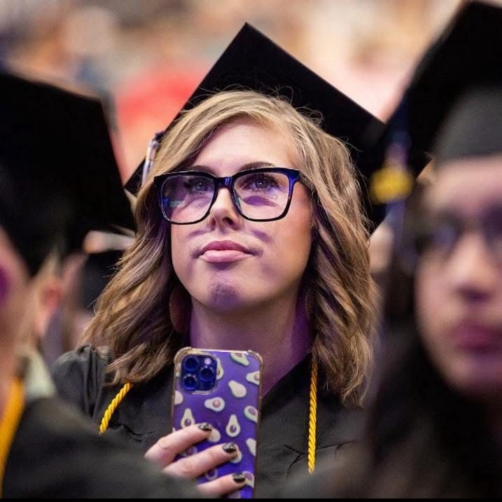 Woman at graduation wearing her cap and gown looks attentively toward stage. She is framed in the foreground by two other graduates out of focus.