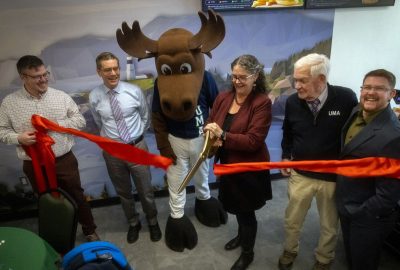 University of Maine at Augusta leadership and mascot Augustus the moose watch with excitement as UMA President Jenifer Cushman cuts a long red ribbon with large ceremonial scissors.