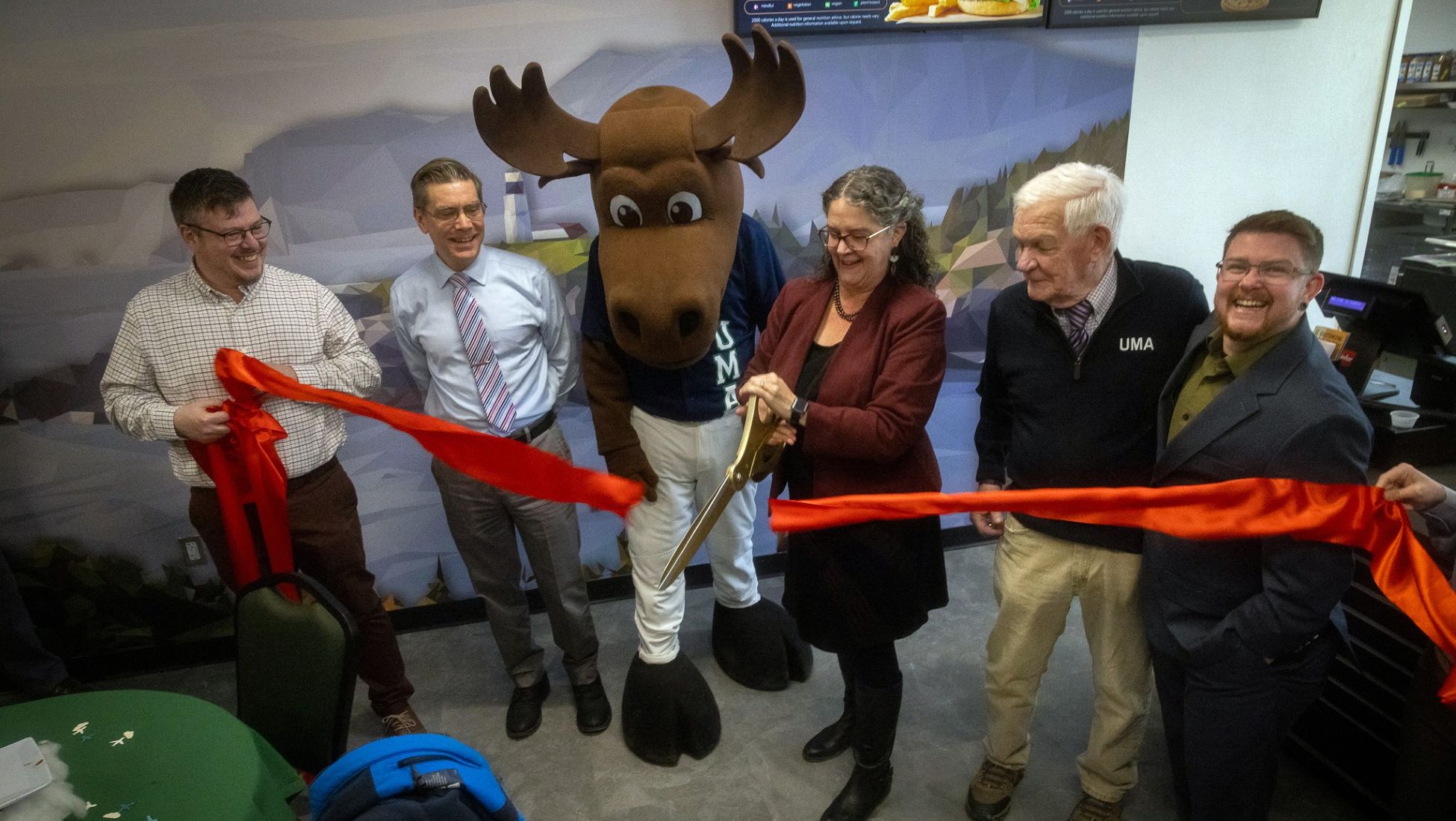 University of Maine at Augusta leadership and mascot Augustus the moose watch with excitement as UMA President Jenifer Cushman cuts a long red ribbon with large ceremonial scissors.
