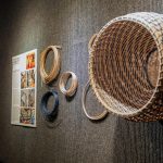 Woven basketry display mounted on a gray gallery wall, featuring a large finished basket and several coils of natural and dyed reed alongside an informational panel.
