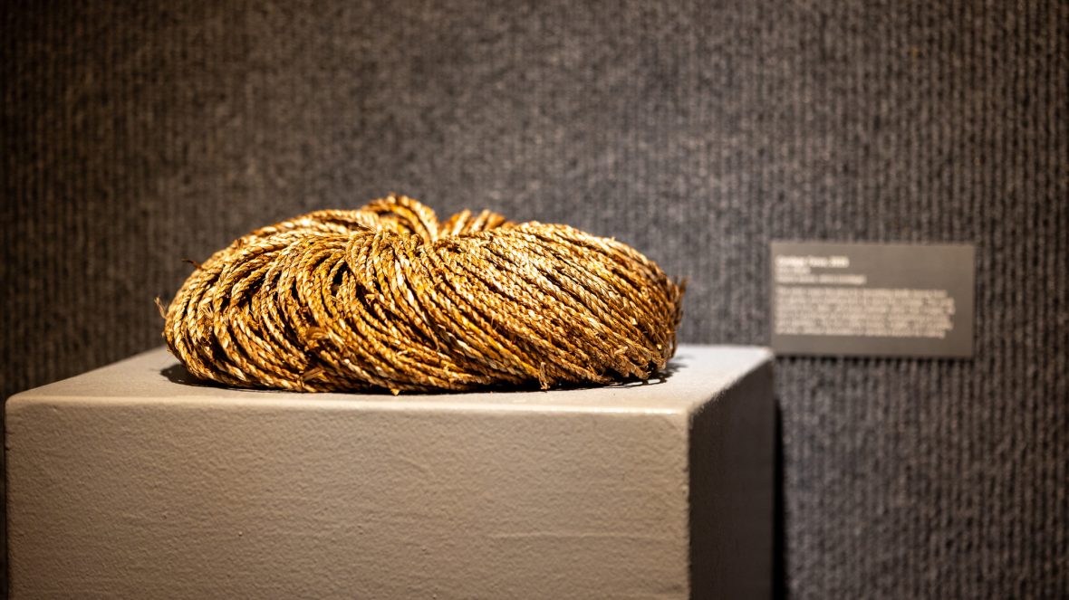Coiled bundle of braided natural fiber resting on a pedestal in a gallery space, with a small exhibit label mounted on the wall behind it.