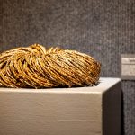 Coiled bundle of braided natural fiber resting on a pedestal in a gallery space, with a small exhibit label mounted on the wall behind it.