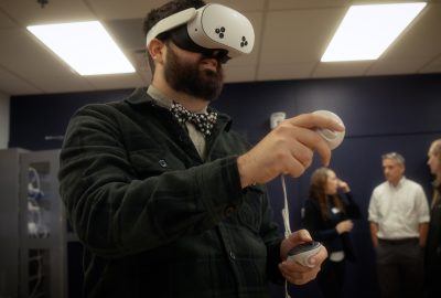 Person wearing a virtual reality headset uses handheld controllers during a VR demonstration in a classroom, with several people talking in the background.