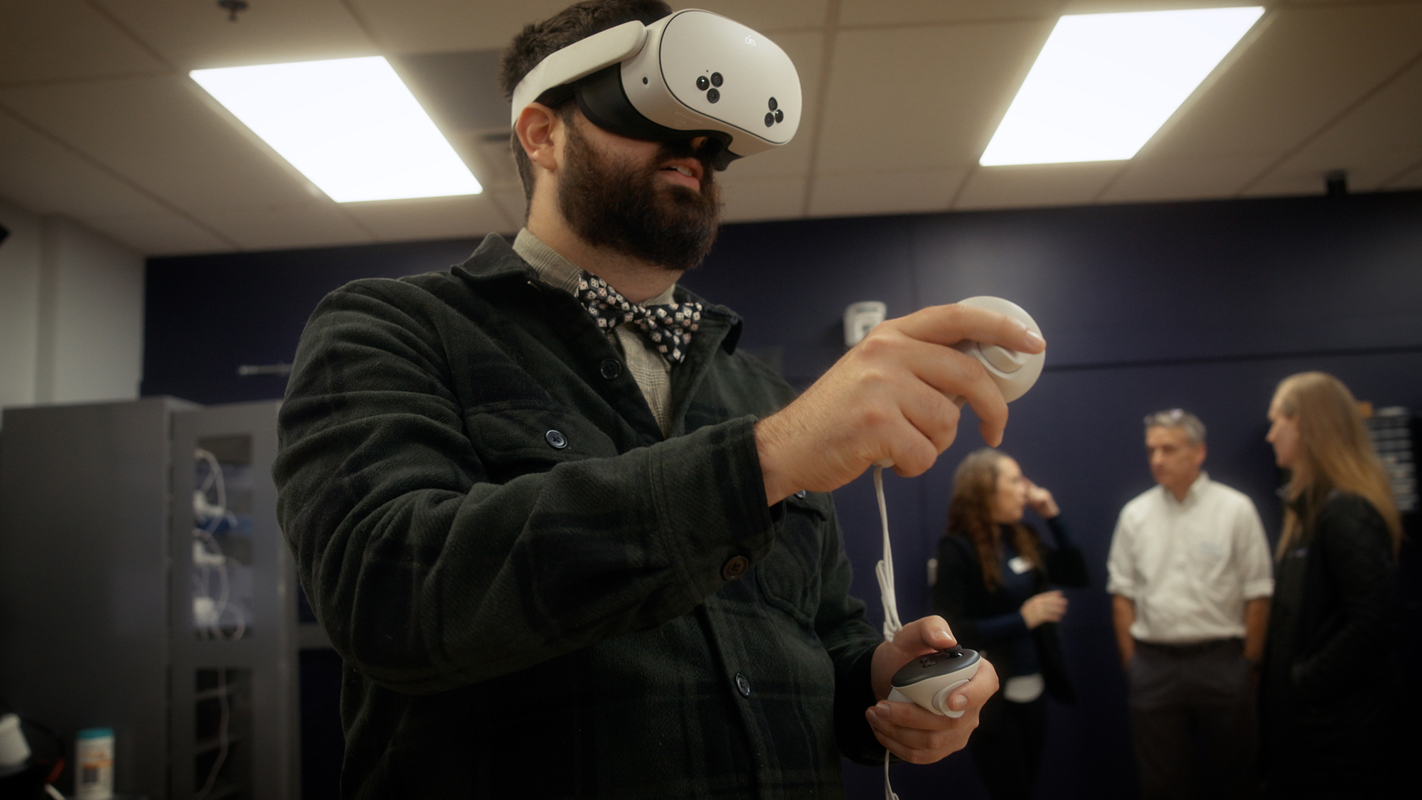 Person wearing a virtual reality headset uses handheld controllers during a VR demonstration in a classroom, with several people talking in the background.