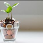 Small green seedling sprouting from a clear glass cup filled with assorted coins, set against a soft, neutral background—symbolizing financial growth or savings.