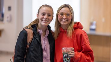 Two UMA students stand together in a campus hallway, smiling at the camera; one wears a backpack and jacket, while the other in a red hoodie holds a clear iced drink with an Aroma Joe’s logo.