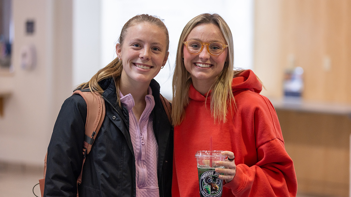Two UMA students stand together in a campus hallway, smiling at the camera; one wears a backpack and jacket, while the other in a red hoodie holds a clear iced drink with an Aroma Joe’s logo.