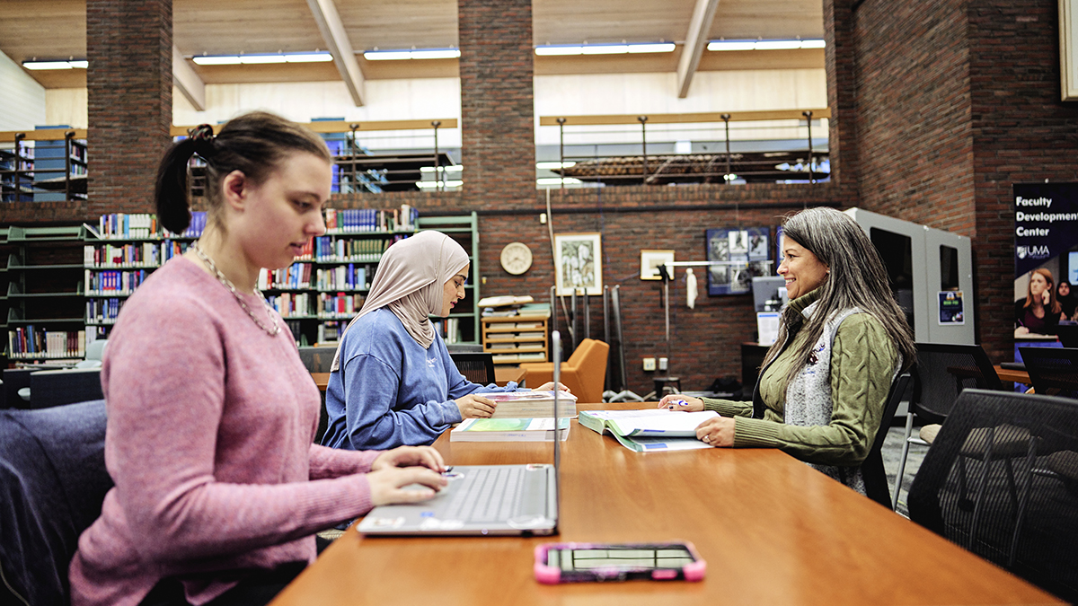 Three people sit at a large table in a campus library; two students work on a laptop and textbooks while a staff member across the table reviews materials with them, with bookshelves and brick walls visible in the background.