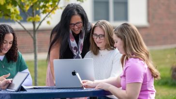 Staff member and three students gather around a laptop and tablet at an outdoor campus table, smiling and working together.