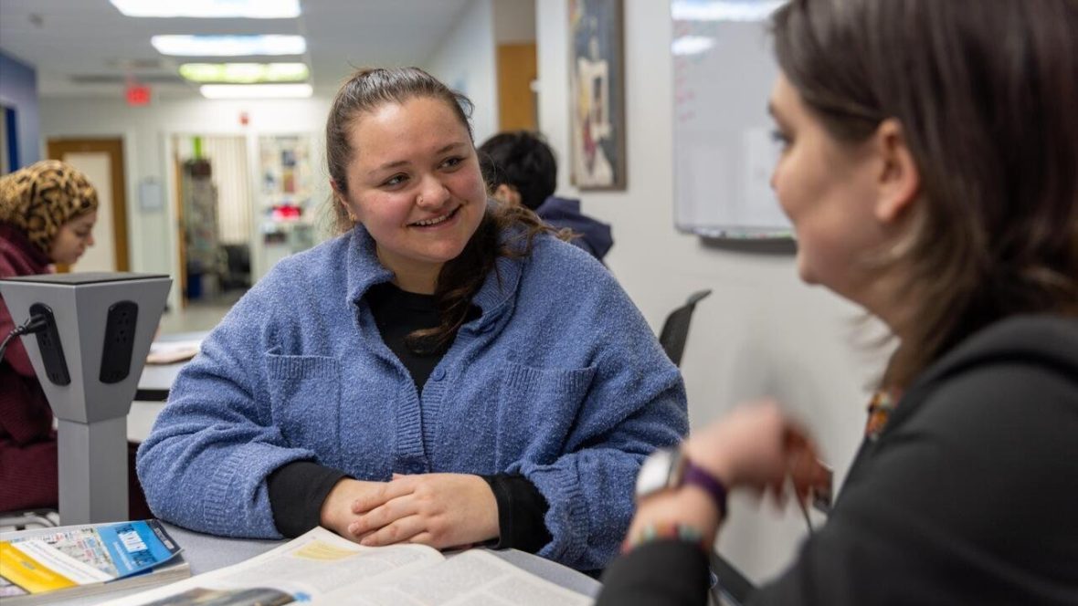 Student in a blue fleece sits at a campus service desk, smiling and talking with a staff member across the counter while paperwork and a card reader sit nearby; another student works at a computer in the background of the hallway office.
