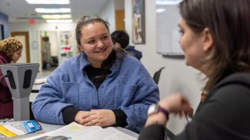 Student in a blue fleece sits at a campus service desk, smiling and talking with a staff member across the counter while paperwork and a card reader sit nearby; another student works at a computer in the background of the hallway office.