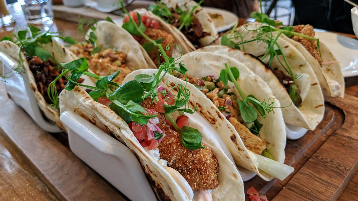 A row of soft tacos arranged in a wooden holder, filled with crispy breaded fish or protein, fresh pico de gallo, greens, and garnished with microgreens, served on a wooden table in a restaurant setting.