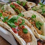 A row of soft tacos arranged in a wooden holder, filled with crispy breaded fish or protein, fresh pico de gallo, greens, and garnished with microgreens, served on a wooden table in a restaurant setting.