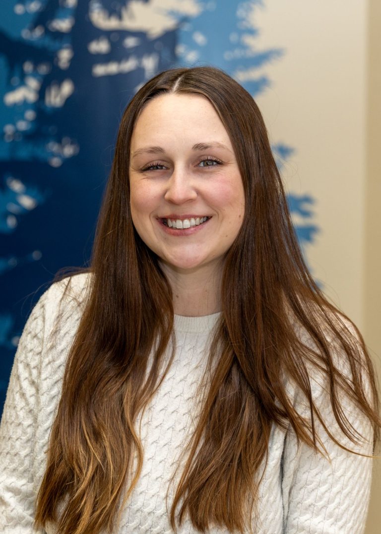 A woman with long hair and a white sweater smiles in front of a wall painted with blue trees on a beige background