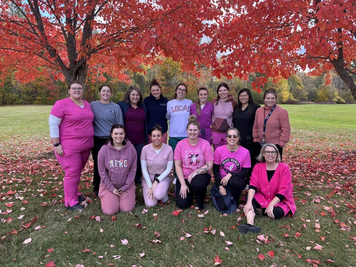 Group of students and faculty wearing pink for Breast Cancer Awareness Month standing together beneath a bright red autumn tree on a grassy lawn.
