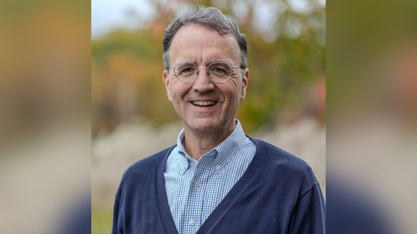 Chris Devlin wearing glasses and a v-necked sweater over a button-up shirt. Out of focus trees in the background.