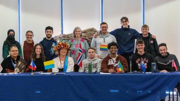 Students and staff pose together behind a table with international flags at a campus cultural celebration.