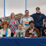 Students and staff pose together behind a table with international flags at a campus cultural celebration.