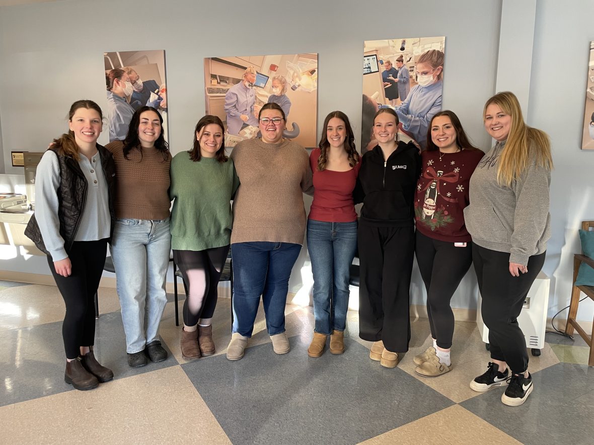 Group of dental students standing together indoors in front of wall photos showing dental procedures and clinical training.