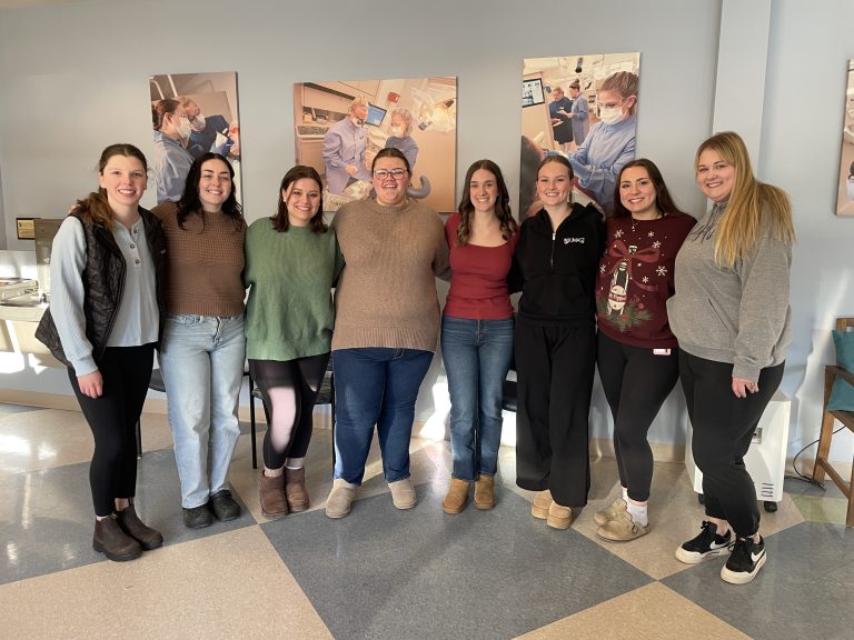 Group of dental students standing together indoors in front of wall photos showing dental procedures and clinical training.