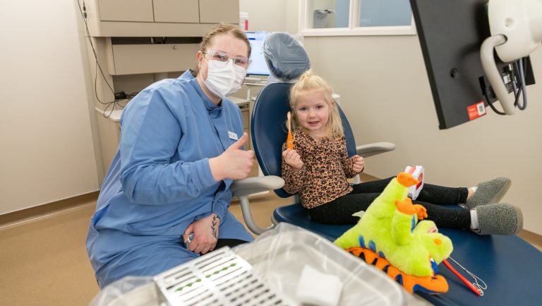 A dental hygiene student wearing a medical mask and blue lab coat gives a thumbs up while posing with her young patient who smiles while holding a tooth brush