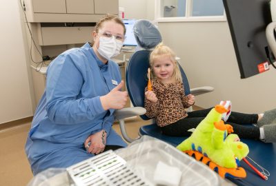 A dental hygiene student wearing a medical mask and blue lab coat gives a thumbs up while posing with her young patient who smiles while holding a tooth brush
