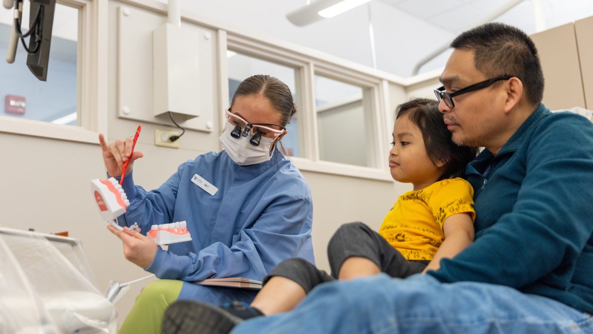 A young girl sits on her father's lap as a dental hygiene student teaches her to care for her teeth