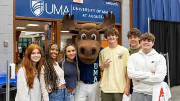 Group of five prospective students smiling with the UMA moose mascot at an event booth under a University of Maine at Augusta sign.
