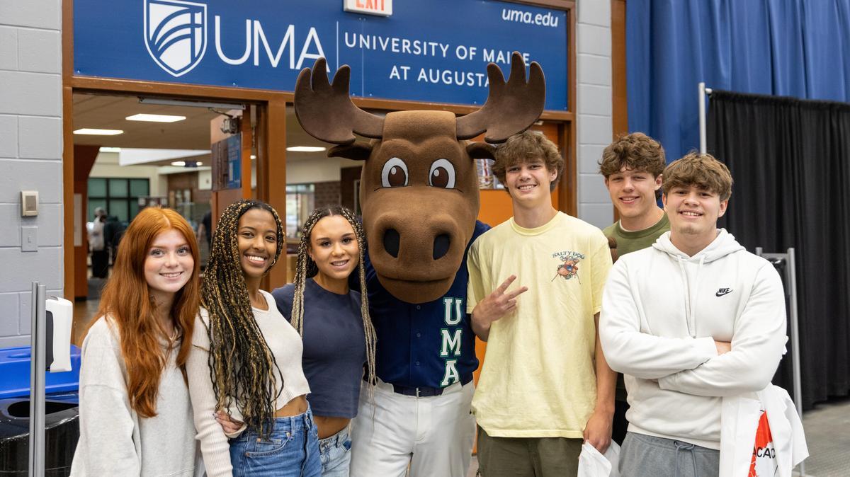 Group of five prospective students smiling with the UMA moose mascot at an event booth under a University of Maine at Augusta sign.