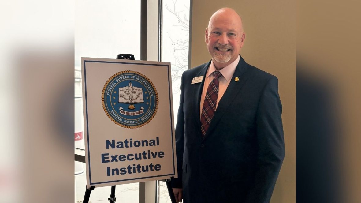 Noel March, wearing a suit and tie, stands beside a sign with a seal that reads, "Federal Bureau of Investigation National Executive Institute"